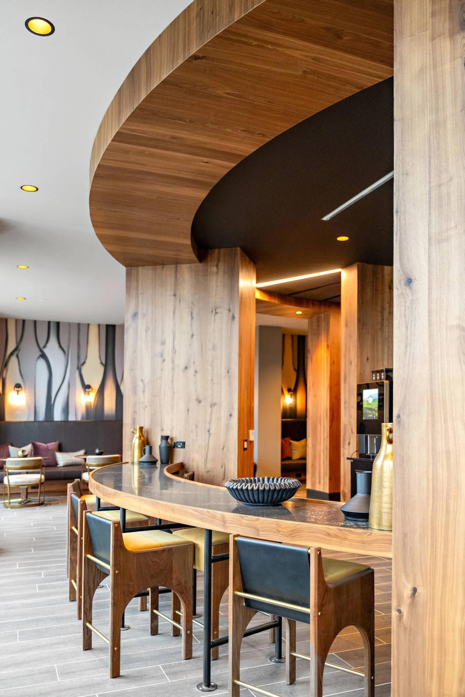 Modern lounge with wooden accents, curved ceiling, high table, and chairs; soft seating in the background.