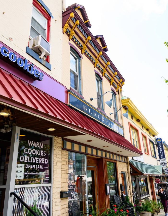 Colorful storefronts, including a cookie shop, line a lively street with trees and outdoor seating.