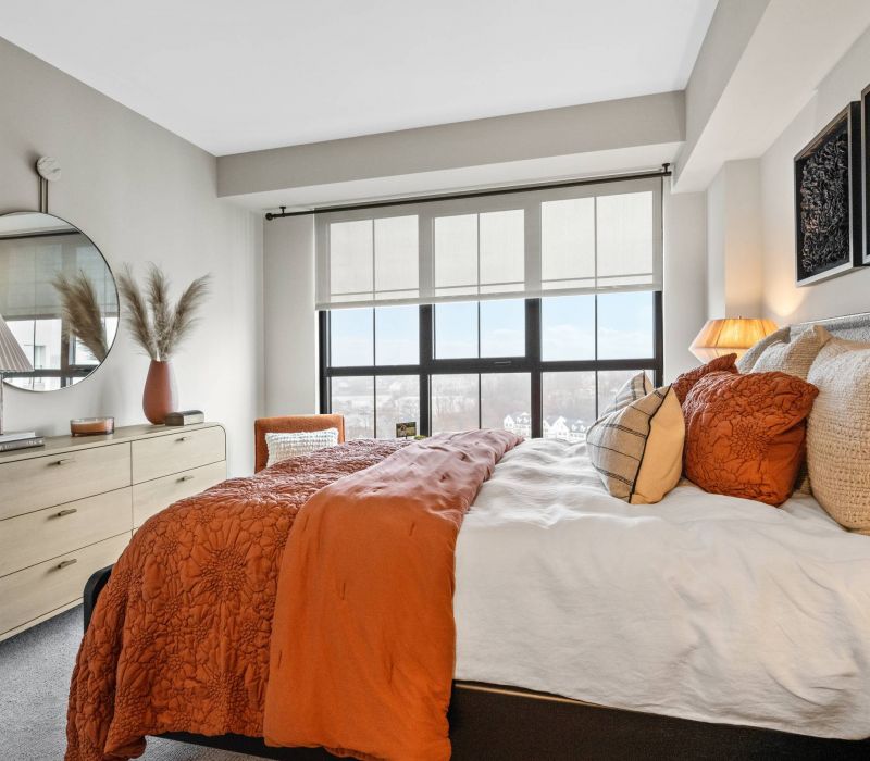 Madison West Elm apartments Modern bedroom with orange and beige bedding, large window, dresser, round mirror, and decorative accents.