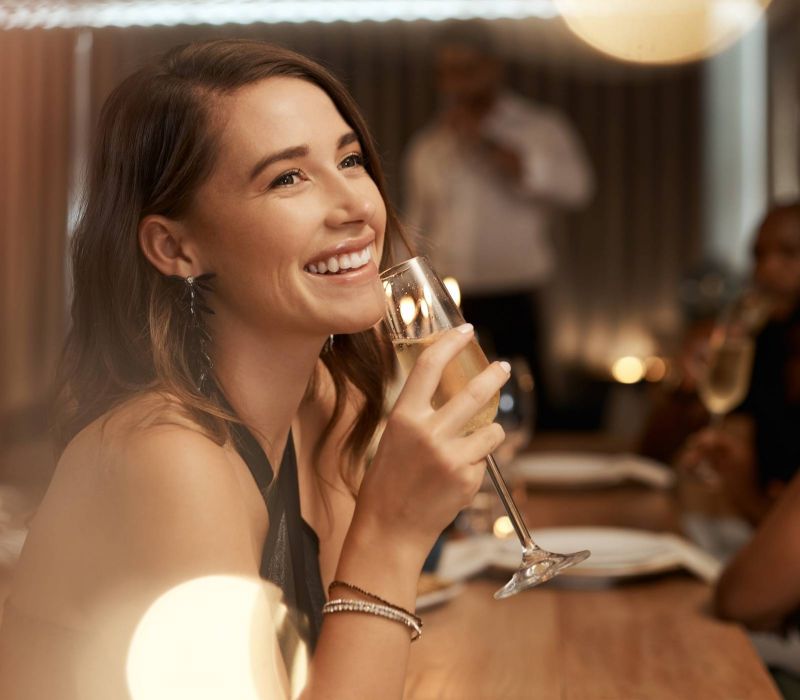 Smiling woman holding a glass of champagne at a restaurant table with people in the background.