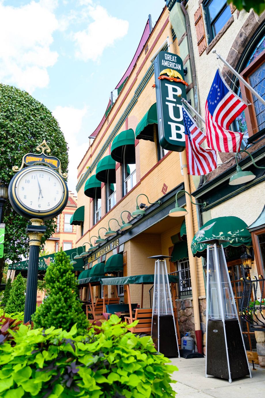 Outdoor view of Great American Pub with green awnings, two American flags, and a vintage street clock in Conshohocken, PA near Madison West Elm apartments.