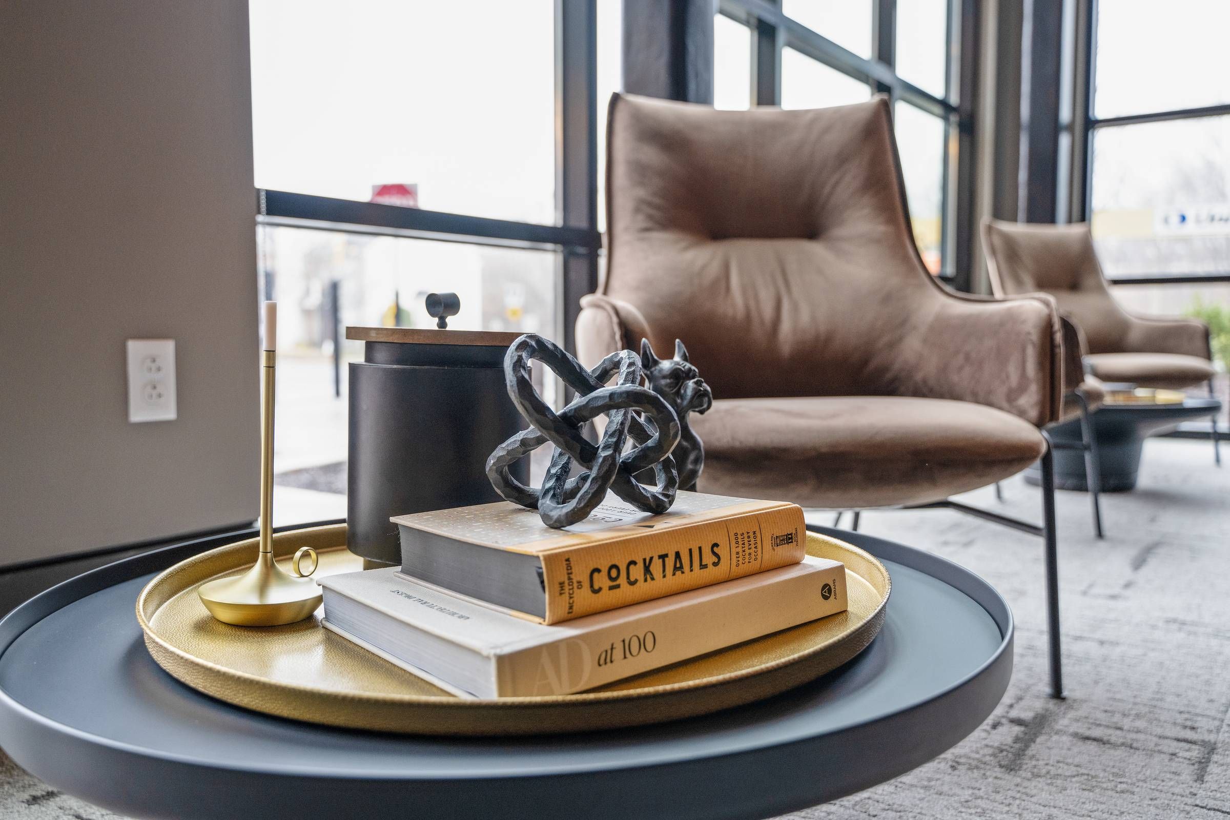 Madison West Elm apartments Modern lounge with brown armchairs, books, and decor on a round coffee table near large windows.