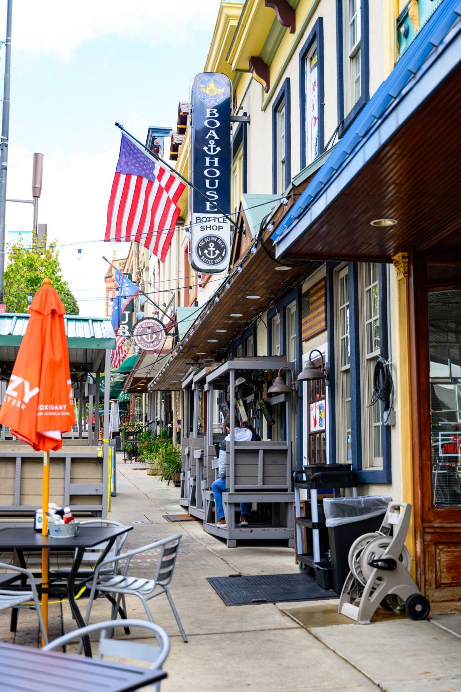 Colorful sidewalk with outdoor seating, American flag, and barber shop sign along a row of shops in Conshohocken, PA near Madison West Elm apartments.