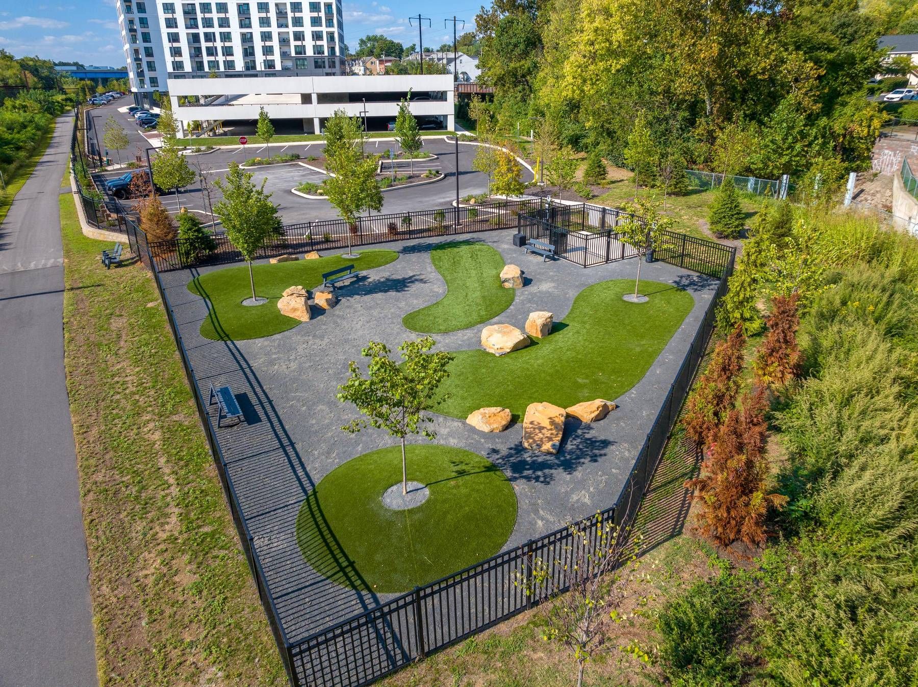 Madison West Elm apartments Aerial view of a fenced dog park with benches, trees, rocks, and winding paths near a parking lot.