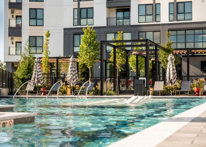 Madison West Elm apartments Modern apartment building with a pool, lounge chairs, umbrellas, and green plants on a sunny day.